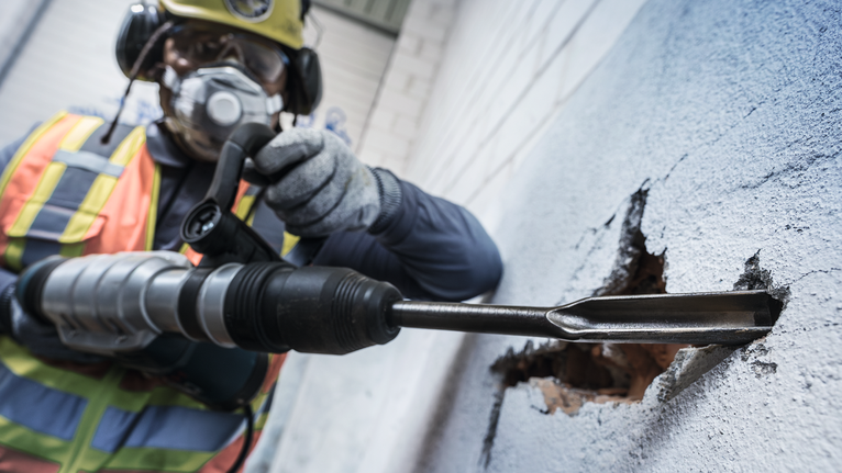 บุคคลที่สวมอุปกรณ์ด้านความปลอดภัยใช้ค้อนโรตารี่เพื่อเจาะผ่านผนังคอนกรีต