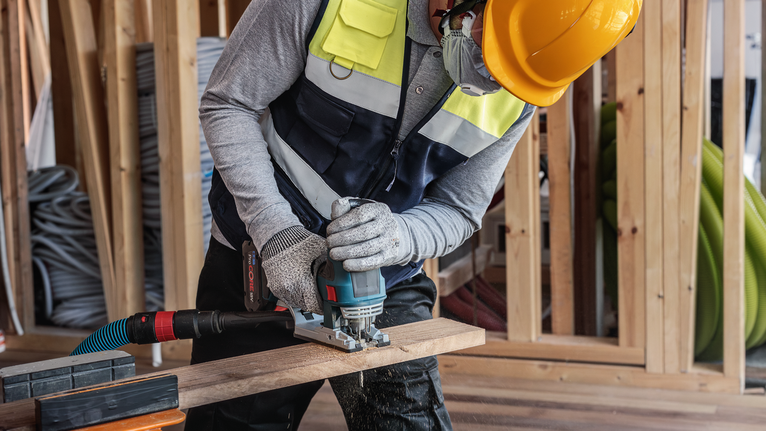 บุคคลที่สวมอุปกรณ์ความปลอดภัยกำลังตัดไม้ด้วยเครื่องมือไฟฟ้าในสถานที่ก่อสร้าง
