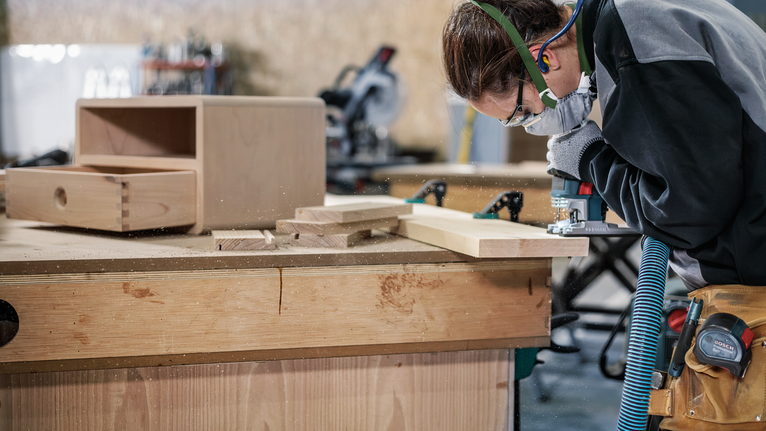 บุคคลที่สวมอุปกรณ์ความปลอดภัยใช้เครื่องมือไฟฟ้าในการขึ้นรูปแผ่นไม้บนโต๊ะทำงาน