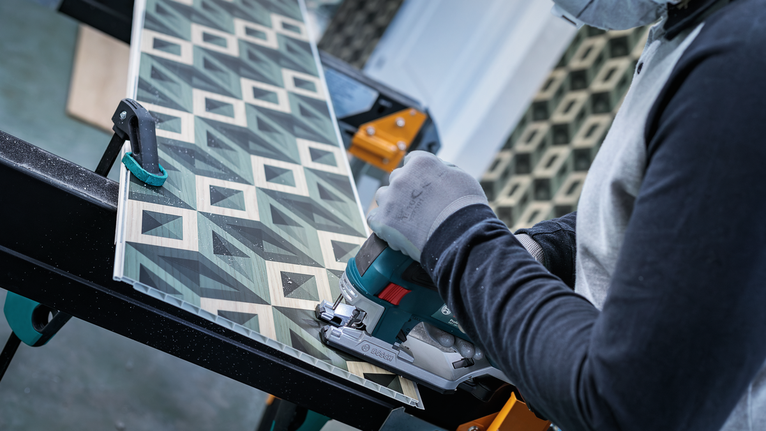 บุคคลที่สวมอุปกรณ์ความปลอดภัยกำลังตัดกระดานที่มีลวดลายด้วยเลื่อยมือถือ