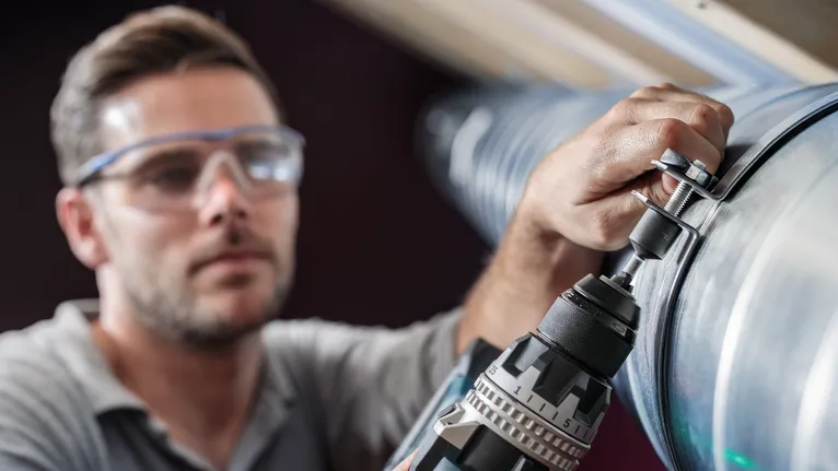 บุคคลที่สวมอุปกรณ์ด้านความปลอดภัยกำลังขันน็อตเข้ากับท่อโลหะด้วยสว่านไฟฟ้า
