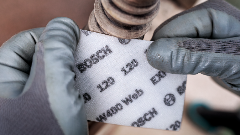 บุคคลที่สวมอุปกรณ์ด้านความปลอดภัยถือแผ่นขัดชนิดหยาบไว้ใกล้กับท่อโลหะ