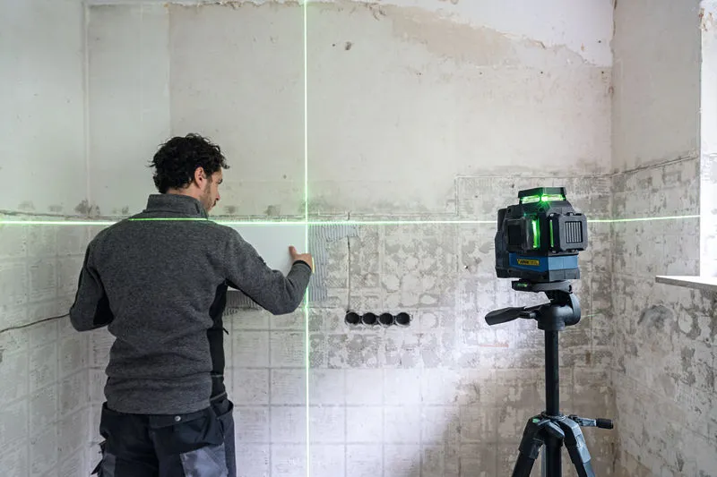 บุคคลกำลังเรียงกระเบื้องบนผนังโดยใช้อุปกรณ์ปรับระดับเลเซอร์สีเขียวบนขาตั้งกล้อง.