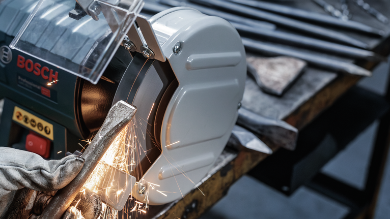 บุคคลที่สวมอุปกรณ์ด้านความปลอดภัยกำลังเจียรเครื่องมือโลหะบนเครื่องเจียรแท่น