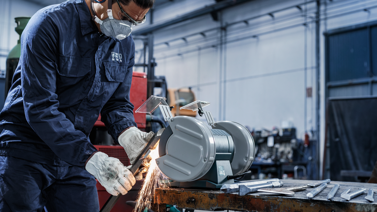 บุคคลที่สวมอุปกรณ์ความปลอดภัยกำลังเจียรโลหะโดยใช้เครื่องเจียรแบบคงที่ในโรงงาน