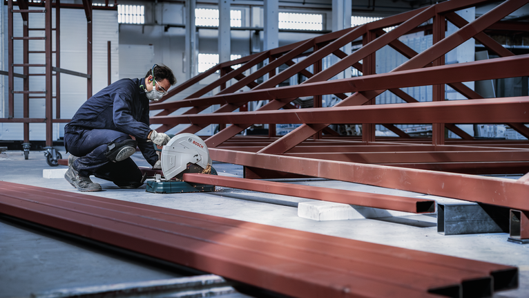 บุคคลที่สวมอุปกรณ์ความปลอดภัยกำลังตัดคานเหล็กด้วยเลื่อยวงเดือนในโรงงานแห่งหนึ่ง