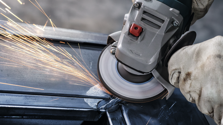 บุคคลที่สวมอุปกรณ์ความปลอดภัยกำลังเจียรคานโลหะด้วยเครื่องเจียรแบบมุม