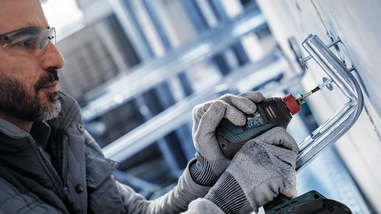 บุคคลที่สวมอุปกรณ์นิรภัยกำลังใช้สว่านไฟฟ้าเพื่อยึดเหล็กยึดเข้ากับผนัง