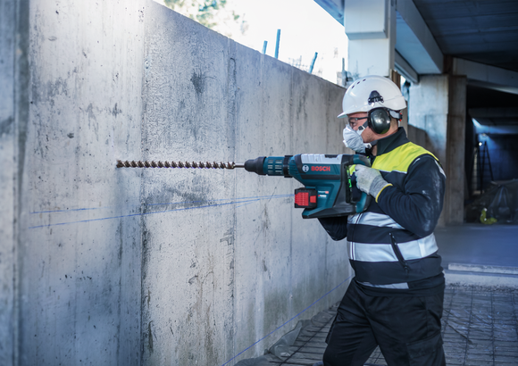 บุคคลที่สวมอุปกรณ์ความปลอดภัยกำลังเจาะเข้าไปในผนังคอนกรีตด้วยค้อนโรตารี่ขนาดใหญ่