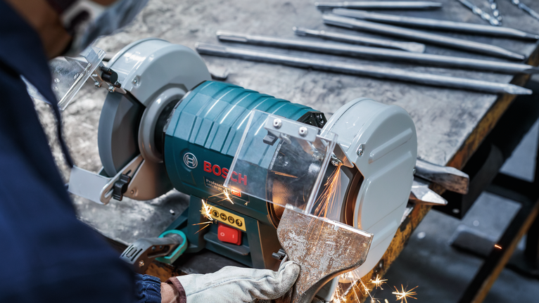 คนงานซึ่งสวมอุปกรณ์ด้านความปลอดภัยกำลังเจียรชิ้นงานโลหะด้วยเครื่องเจียรแบบแท่น.