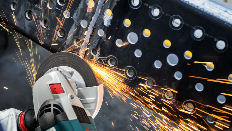 บุคคลที่สวมอุปกรณ์ป้องกันความปลอดภัยกำลังบดคานโลหะด้วยเครื่องมือไฟฟ้า ทำให้เกิดประกายไฟ.