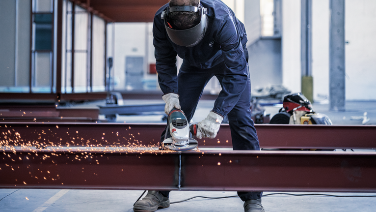 บุคคลที่สวมอุปกรณ์ป้องกันความปลอดภัยกำลังบดคานเหล็กซึ่งก่อให้เกิดประกายไฟสว่าง.