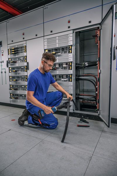 บุคคลที่สวมอุปกรณ์ด้านความปลอดภัยตัดสายเคเบิลหนาที่ด้านหน้าแผงไฟฟ้าเปิดอยู่.