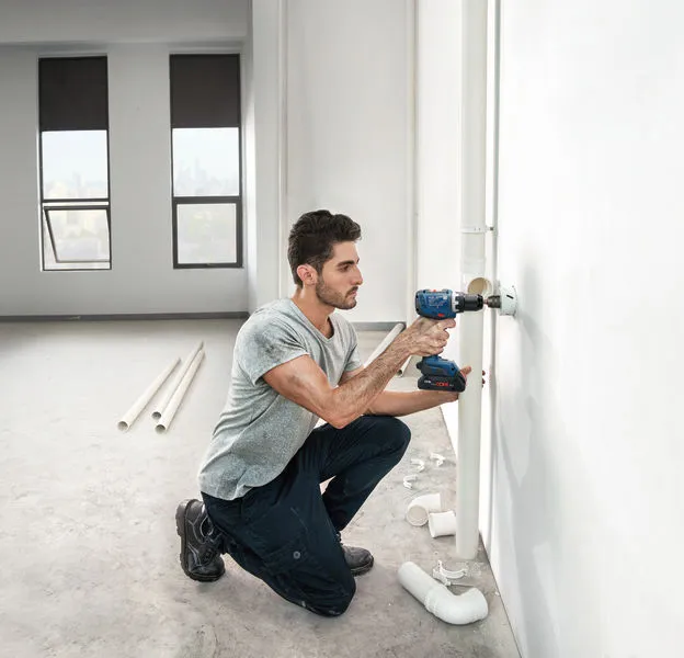 คนกำลังเจาะเข้าไปในผนังข้างท่อประปาในห้องที่ตกแต่งเสร็จเพียงบางส่วน.