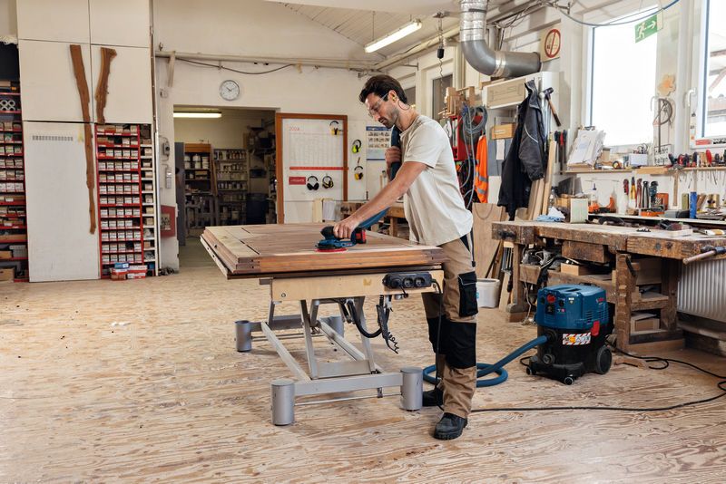 บุคคลที่สวมอุปกรณ์ด้านความปลอดภัยกำลังขัดพื้นไม้ด้วยเครื่องขัดแบบสุ่มวงโคจรแบบไร้สาย.