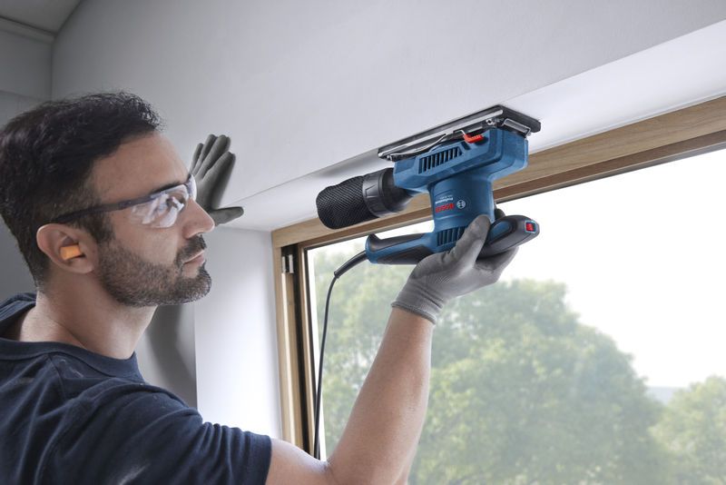 บุคคลที่สวมอุปกรณ์ด้านความปลอดภัยกำลังขัดกรอบหน้าต่างด้วยเครื่องขัดแบบสั่น.