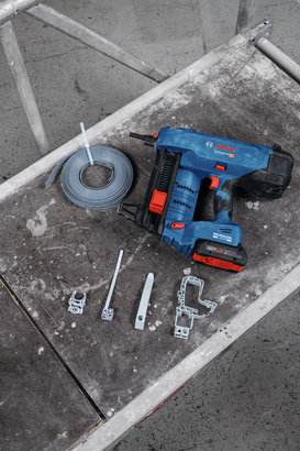 Object #200485878: GNB_18V-40_Application_Packshot_4629 ปืนยิงตะปูไร้สายและตัวยึดโลหะวางเรียงกันบนโต๊ะทำงานที่เต็มไปด้วยฝุ่นละออง