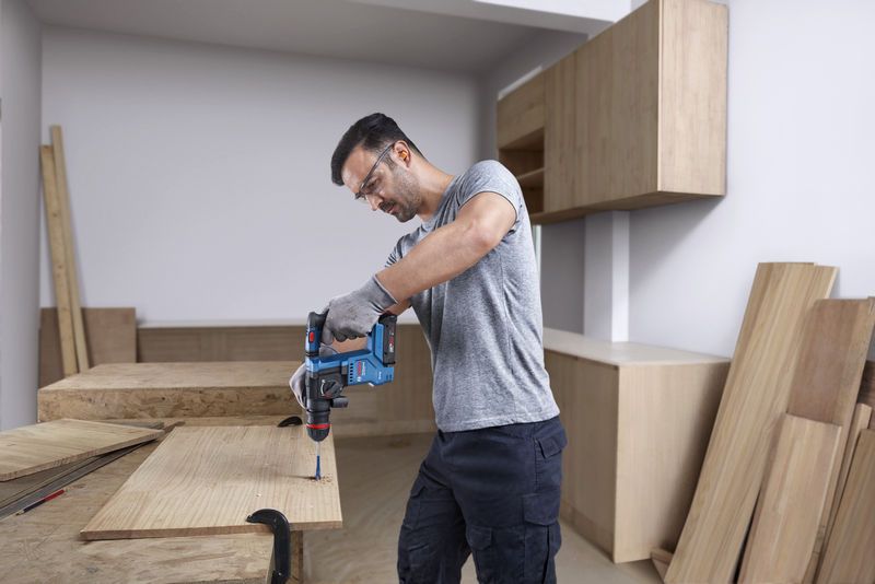 บุคคลที่สวมอุปกรณ์ด้านความปลอดภัยกำลังเจาะเข้าไปในไม้โดยใช้ค้อนโรตารี่ไร้สาย.