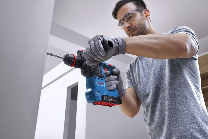 บุคคลที่สวมอุปกรณ์ด้านความปลอดภัยกำลังเจาะเข้าไปในผนังคอนกรีตโดยใช้สว่านโรตารี่ไร้สาย.