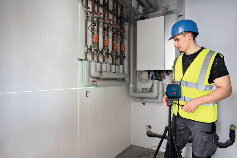 บุคคลที่สวมอุปกรณ์ด้านความปลอดภัยกำลังตั้งอุปกรณ์วัดระดับเลเซอร์ไว้ใกล้ท่อบนผนัง.