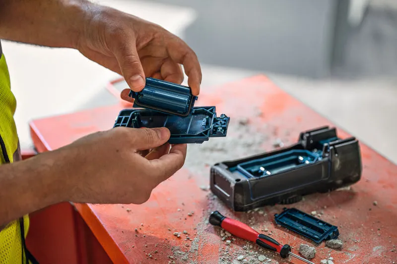 บุคคลที่สวมอุปกรณ์ด้านความปลอดภัยกำลังเปลี่ยนแบตเตอรี่ในเครื่องมือไฟฟ้าบนโต๊ะทำงานที่เต็มไปด้วยฝุ่นละออง.