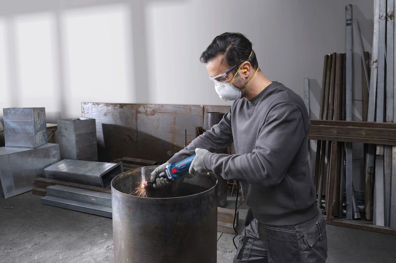 บุคคลที่สวมอุปกรณ์ป้องกันความปลอดภัยกำลังเจียรขอบถังโลหะด้วยเครื่องเจียรตรง.