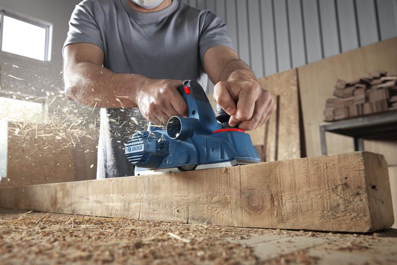 บุคคลที่สวมอุปกรณ์ป้องกันความปลอดภัยกำลังไสไม้คานและมีเศษไม้กระเด็นออกมา.