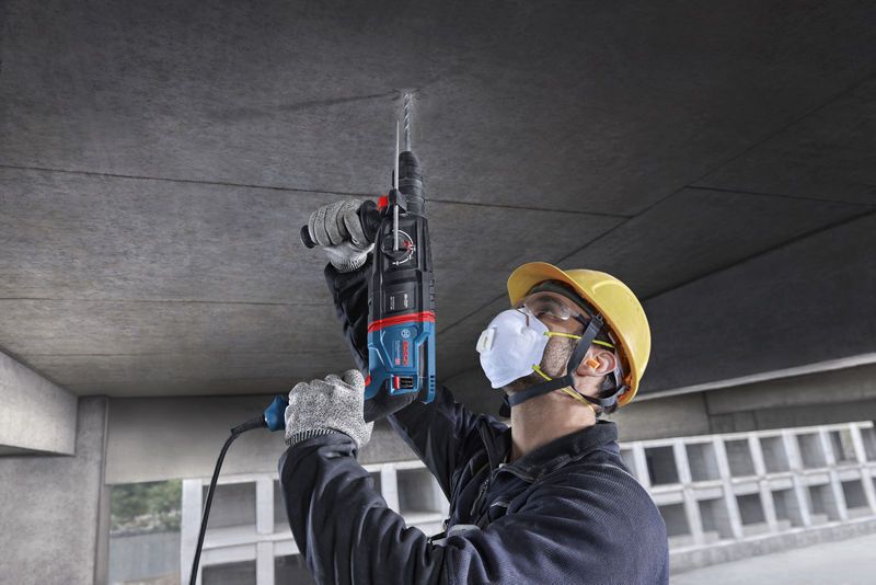 บุคคลที่สวมอุปกรณ์ด้านความปลอดภัยกำลังเจาะเข้าไปในเพดานคอนกรีตโดยใช้ค้อนโรตารี่.