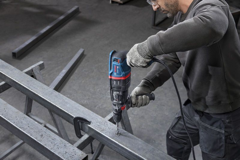 บุคคลที่สวมอุปกรณ์ด้านความปลอดภัยกำลังเจาะเข้าไปในคานโลหะด้วยค้อนโรตารี่.