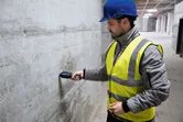 บุคคลที่สวมอุปกรณ์ความปลอดภัยตรวจสอบความชื้นของผนังคอนกรีตด้วยเครื่องวัดความชื้นของวัสดุ.
