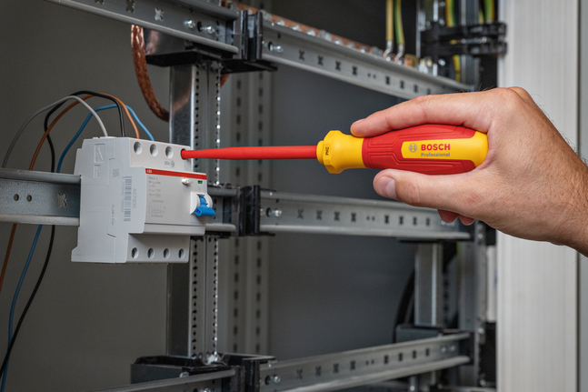 บุคคลกำลังใช้ไขควงหุ้มฉนวนปรับแต่งสายไฟในแผงควบคุมไฟฟ้า