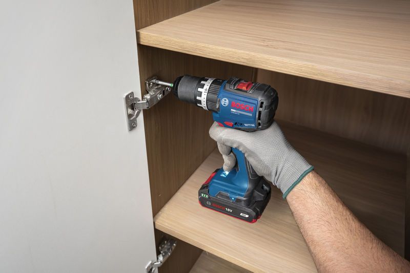 บุคคลที่สวมอุปกรณ์ป้องกันความปลอดภัยกำลังใช้สว่านกระแทกไร้สายเพื่อติดตั้งบานพับตู้