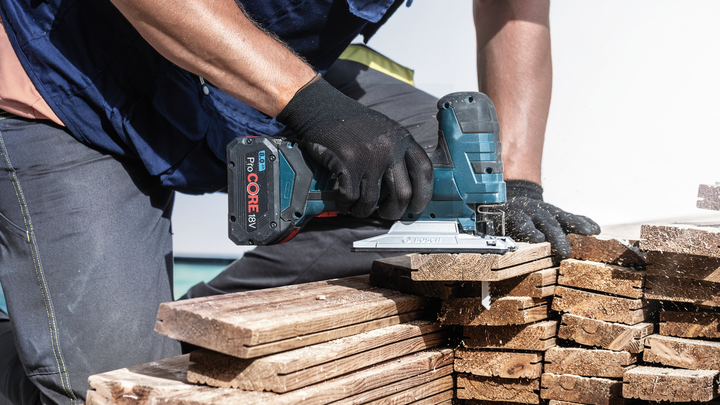 บุคคลที่สวมอุปกรณ์ความปลอดภัยใช้จิ๊กซอว์ไร้สายตัดไม้กระดานที่วางซ้อนกัน.