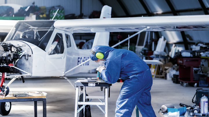 บุคคลที่สวมอุปกรณ์ด้านความปลอดภัยกำลังเจาะชิ้นส่วนโลหะบนโต๊ะทำงานใกล้กับเครื่องบินขนาดเล็ก.