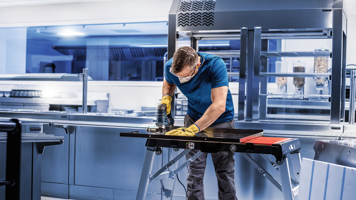 บุคคลที่สวมอุปกรณ์ด้านความปลอดภัยใช้เครื่องมือไฟฟ้าตัดโลหะในห้องครัวเชิงพาณิชย์.