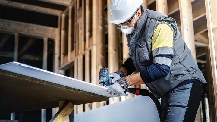 บุคคลที่สวมอุปกรณ์ความปลอดภัยใช้สว่านไฟฟ้าเจาะแผ่นยิปซัมในโครงอาคาร.