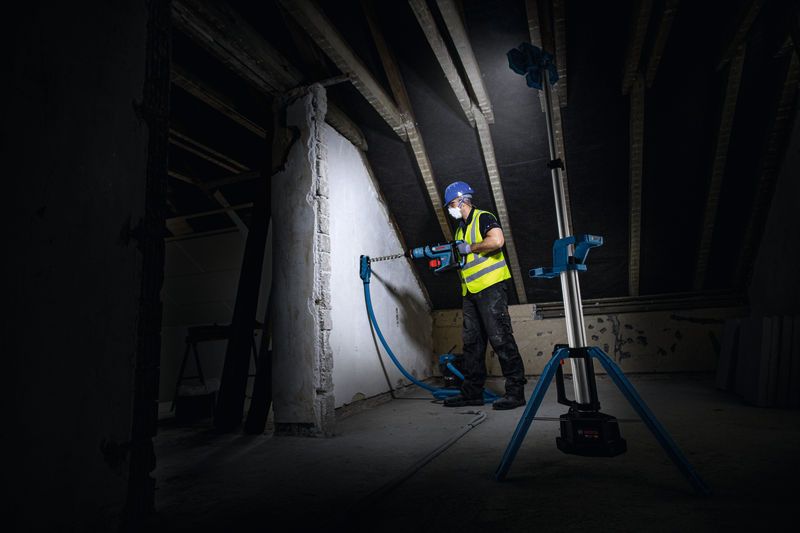 บุคคลที่สวมอุปกรณ์ด้านความปลอดภัยกำลังเจาะเข้าไปในผนังซึ่งมีแสงสว่างจากไฟส่องสว่างหน้างานแบบไร้สาย