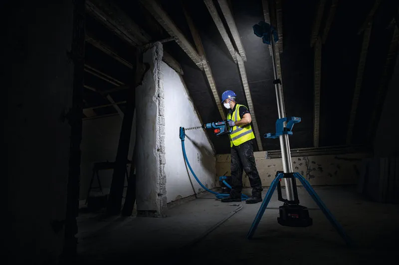 บุคคลที่สวมอุปกรณ์ด้านความปลอดภัยกำลังเจาะเข้าไปในผนังซึ่งมีแสงสว่างจากไฟส่องสว่างหน้างานแบบไร้สาย