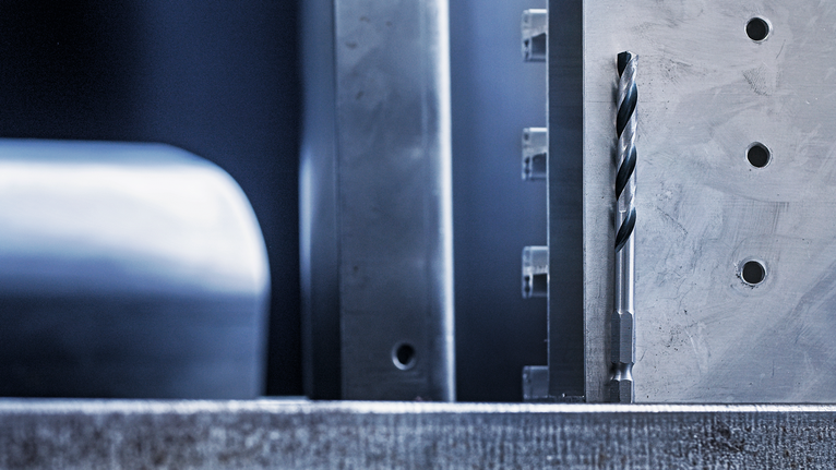 ดอกสว่านโลหะวางเรียงแนวตั้งข้าง ๆ พื้นผิวโลหะที่ผ่านการกลึงซึ่งมีรูเจาะไว้