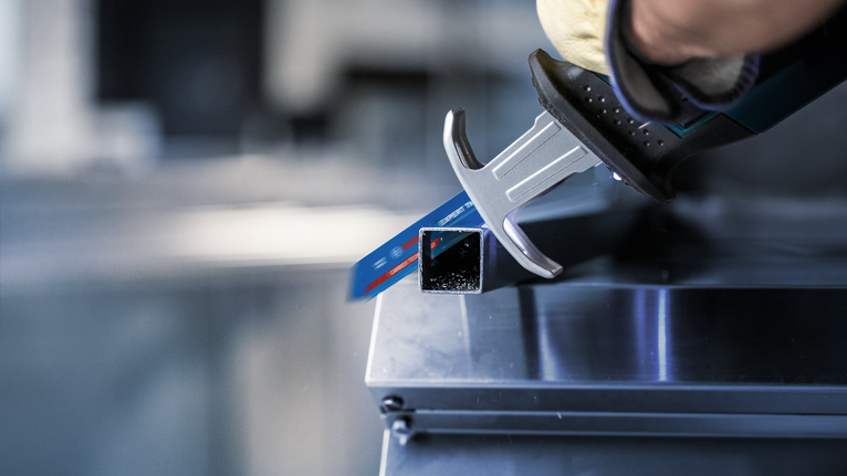 บุคคลที่สวมอุปกรณ์ความปลอดภัยกำลังตัดท่อโลหะทรงสี่เหลี่ยมด้วยเลื่อยไฟฟ้าแบบลูกสูบ