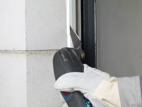 คนงานสวมอุปกรณ์ด้านความปลอดภัยกำลังตัดวัสดุซีลออกจากกรอบหน้าต่างโดยใช้เครื่องมือไฟฟ้า.